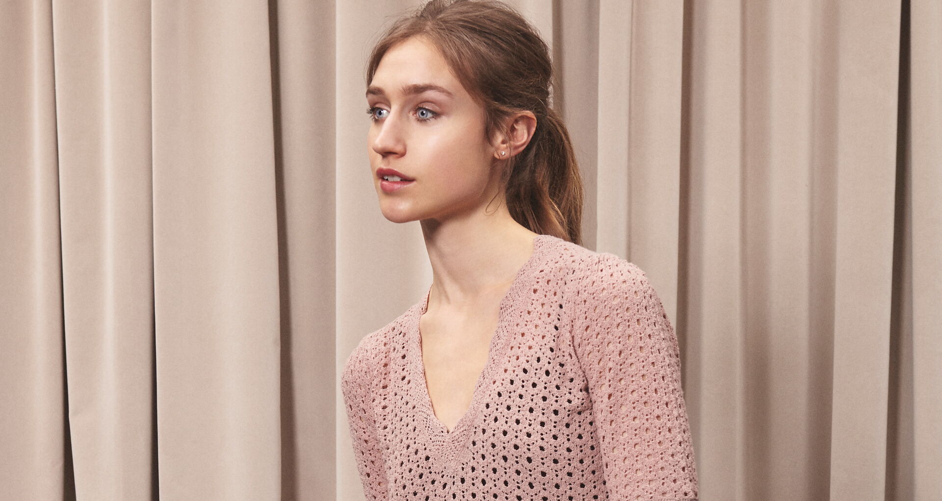 Junge Frau mit Zopf trägt einen Strickpuller mit V-Ausschnitt in der Farbe Alt-Rosé