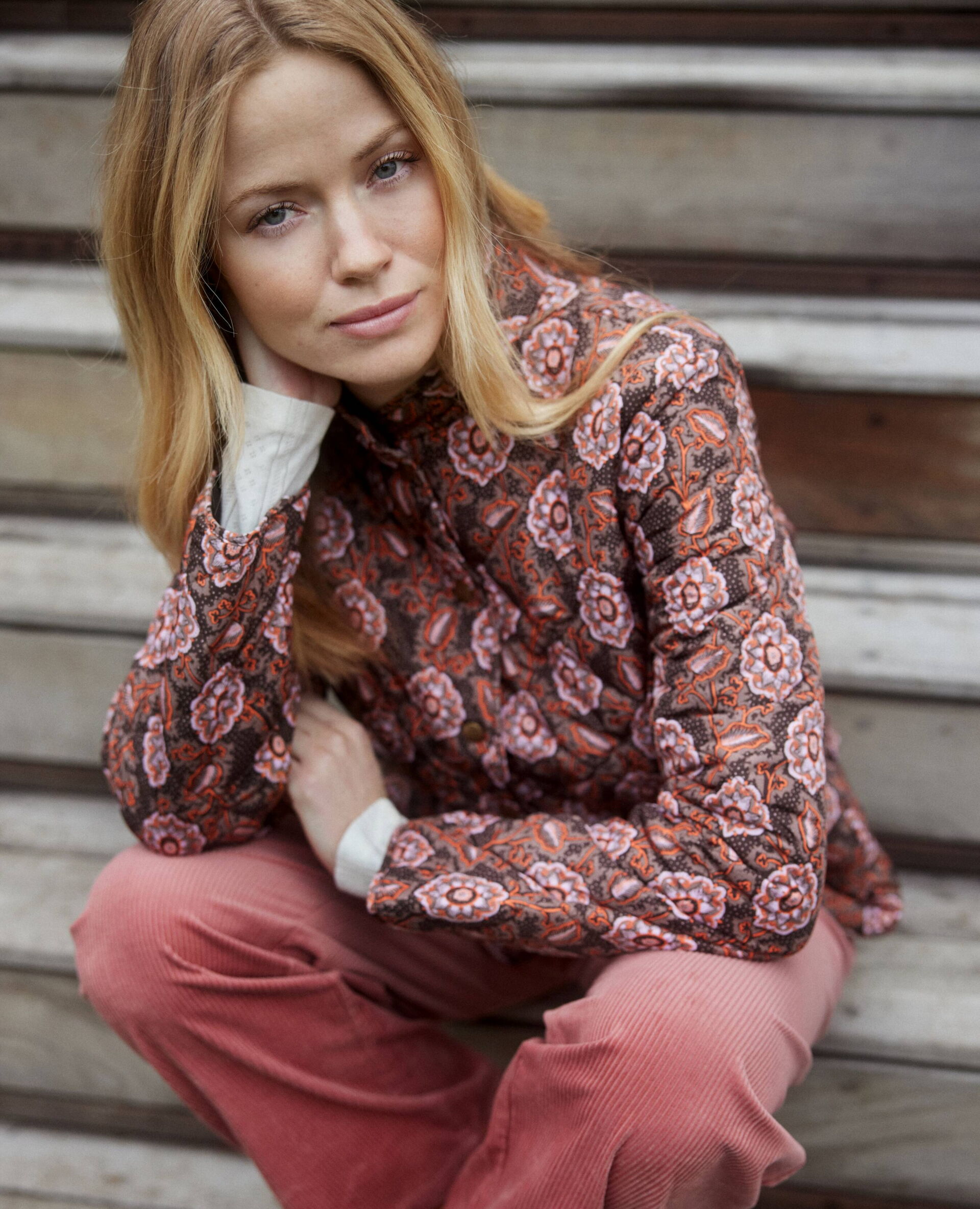 Blonde, junge Frau in einer dunklen, femininen Bluse mit Oll-Over-Print in Rosa-Tönen und Cordhose in Rosa